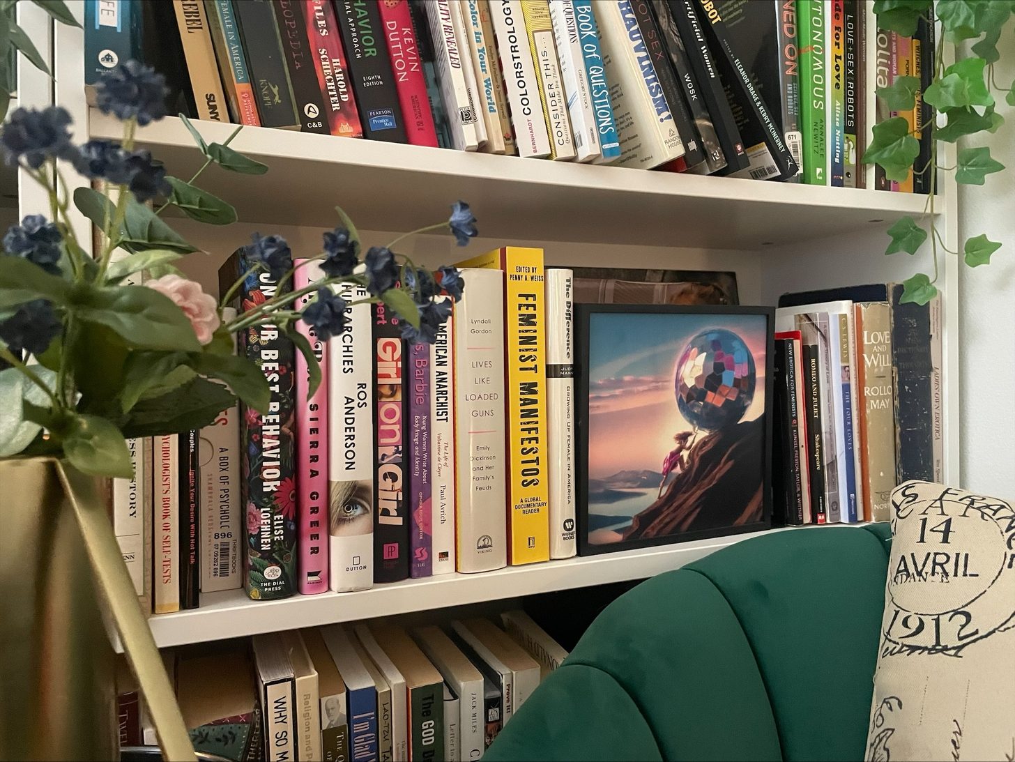 A framed Amor Vincit Absurdum print of a woman with a disco-ball head, displayed on a bookshelf among feminist books beside a green velvet chair